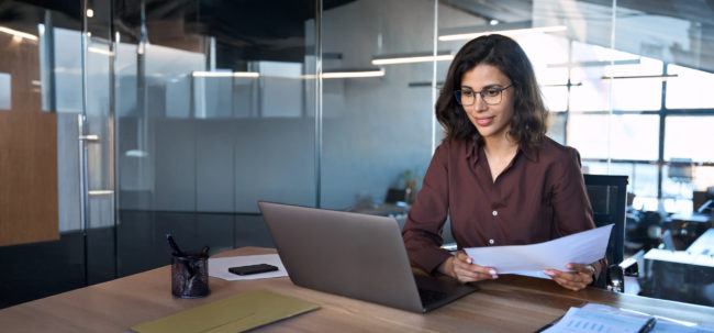 Focused latin hispanic young business woman working on laptop computer reading financial document report in office. Accountant entrepreneur manager businesswoman doing paperwork using pc. Copy space